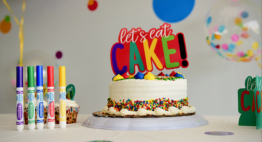 A birthday cake that says "let's eat cake" and Crayola markers sitting on a table.