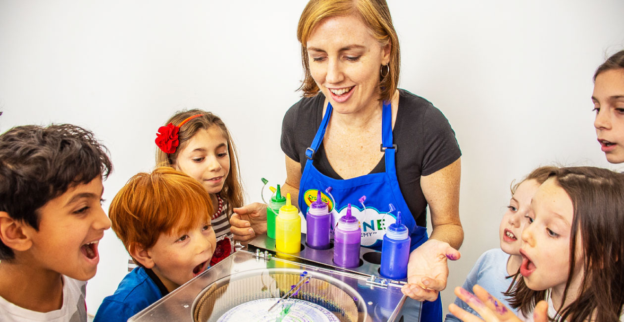 A Crayola Imagine Arts Academy art instructor showing 6 children looking on in amazement how spin art works with different co
